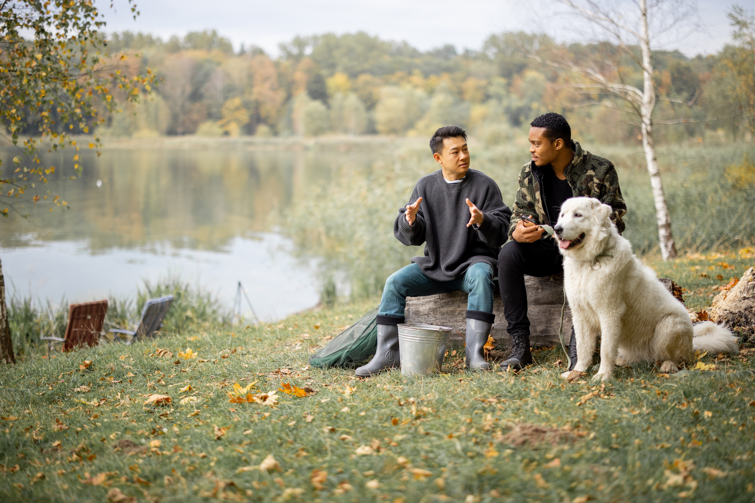 Two men chat by a lake with their dog