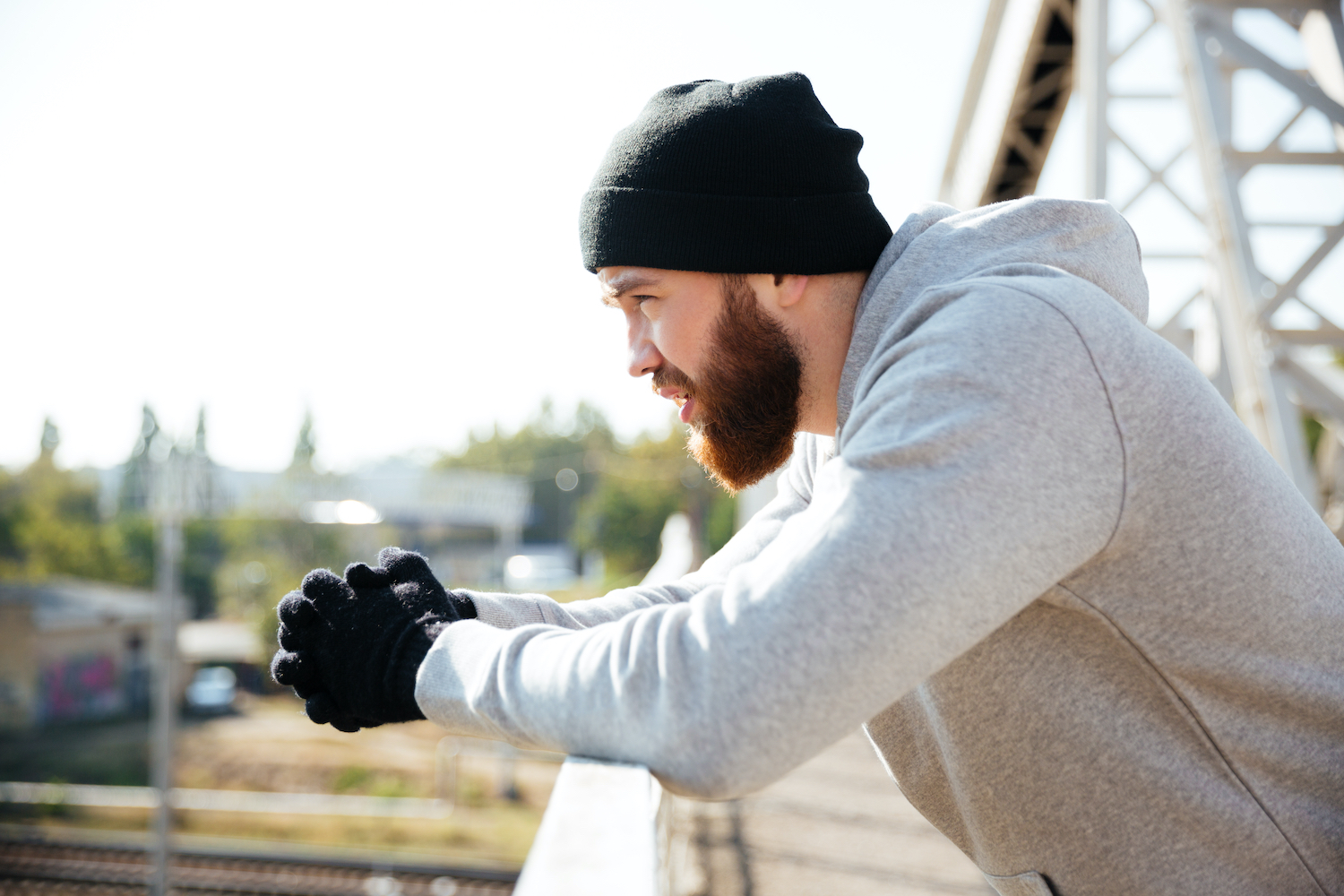 Side view of a young bearded sportsman in hat resting after workout while standing at the bridge