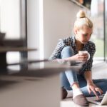 Woman using laptop at home while having coffee sitting on the floor