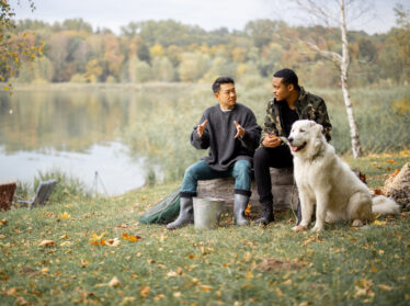 Two men chat by a lake with their dog
