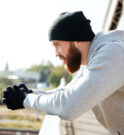 Side view of a young bearded sportsman in hat resting after workout while standing at the bridge